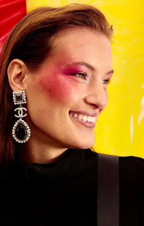 Woman with pink blush and black Chanel earrings smiles against a yellow and red background in an indoor, close-up fashion shot.