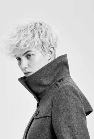 Monochrome portrait of a person with short, textured blonde hair wearing a gray wool coat against a light gray gradient background.