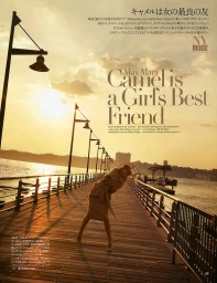 Model wearing a camel coat on a wooden pier during a golden sunset with lampposts lining the waterfront.