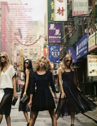 Four female models walking down a busy city street lined with vibrant storefront signs and traditional banners.