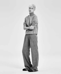Model with short blonde hair wearing a grey turtleneck sweater and trousers against a white background
