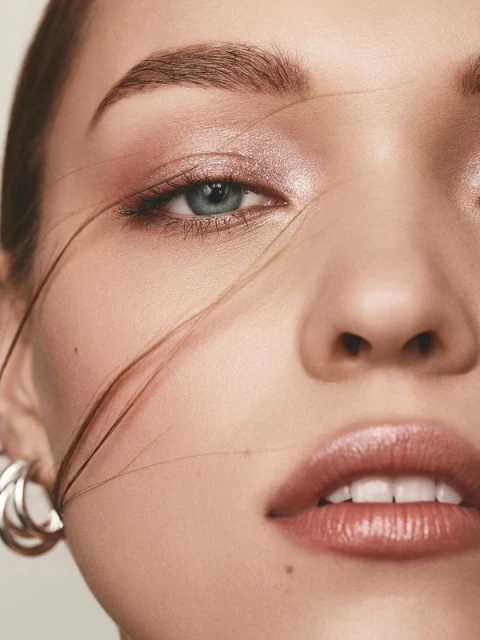 Close-up of a woman's face featuring light makeup, silver hoop earrings, and loose strands of brown hair across her face, against a neutral background.