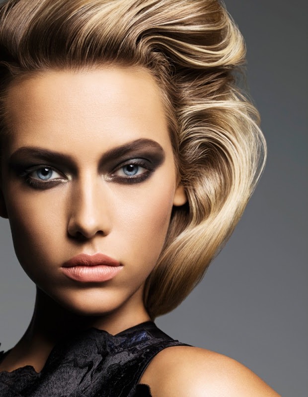 Close-up beauty shot of an adult woman with fair skin, blonde hair styled upwards, and striking black eye makeup, set against a gray background.
