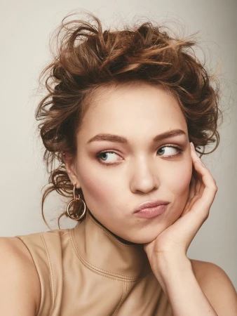 Close-up portrait of a woman with curly hair resting her chin on her hand, wearing a beige high-neck top and gold hoop earrings.