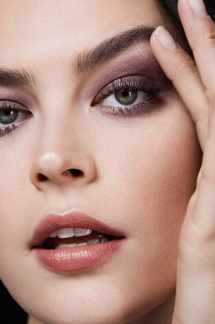Close-up beauty shot of a woman with purple eyeshadow, full lips with lipstick, and a hand near her face showcasing her makeup look.