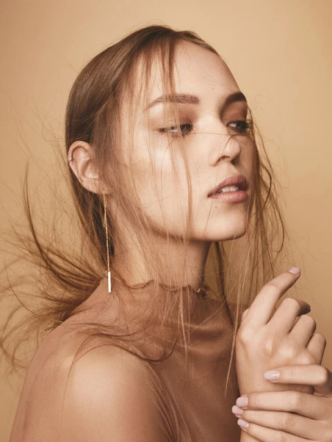 Portrait of a woman with long hair and sheer top, set against a tan background. She is wearing a linear dangle earring.
