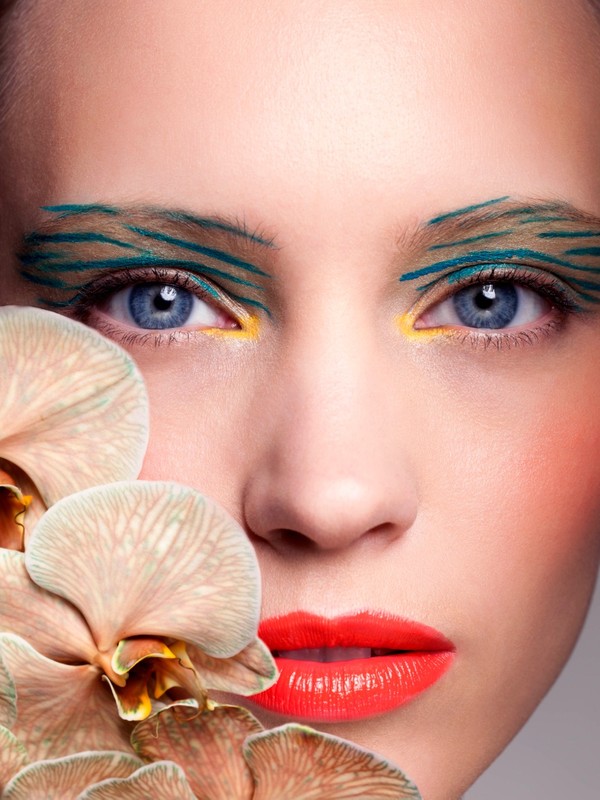 Close-up portrait of a fair-skinned woman with artistic blue and yellow makeup, paired with pale peach-colored orchid blossoms.