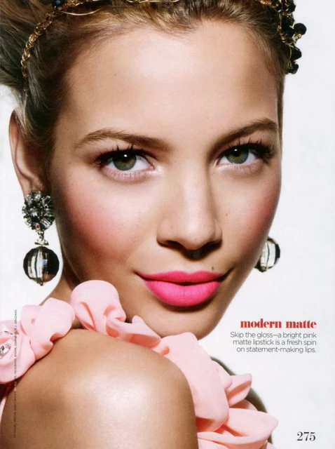 Close-up beauty shot of a blonde woman with pink lipstick, beaded headband, and statement earrings, posing against a bright background.