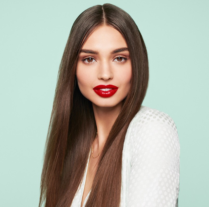 Close-up portrait of a beautiful adult woman with long straight brown hair, red lipstick, wearing a white top against a light blue background.