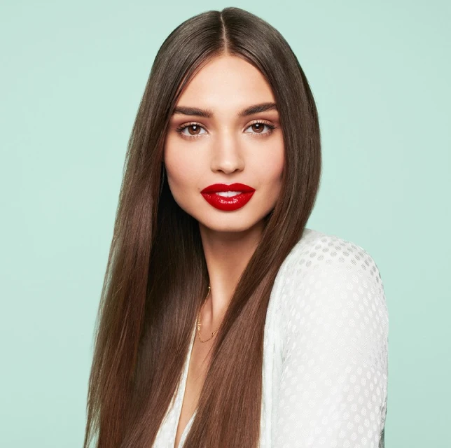 Close-up portrait of a beautiful adult woman with long straight brown hair, red lipstick, wearing a white top against a light blue background.