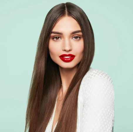 Close-up portrait of a beautiful adult woman with long straight brown hair, red lipstick, wearing a white top against a light blue background.