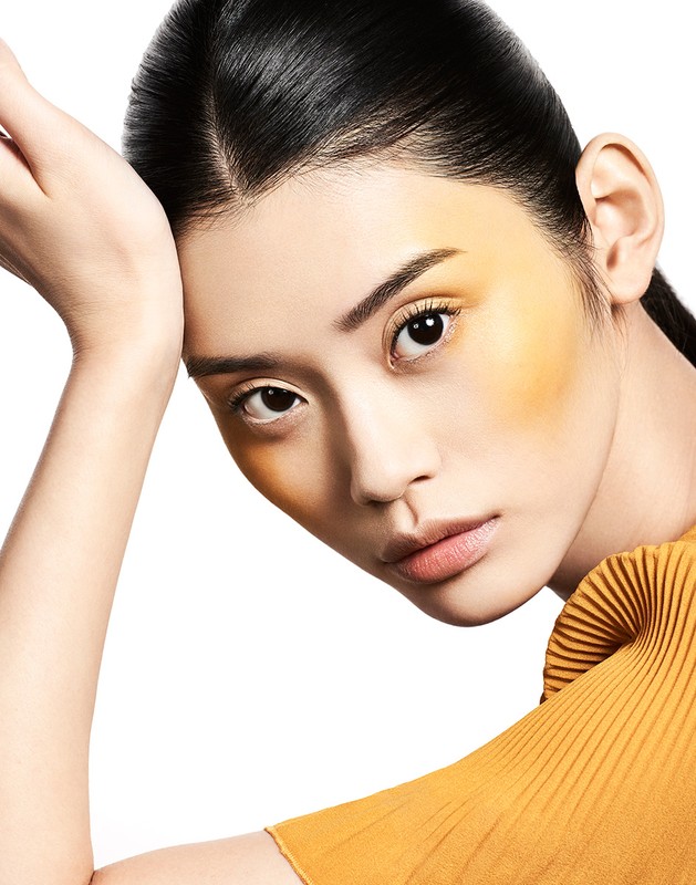 Close-up portrait of a stylish Asian woman with golden blush makeup, wearing a mustard-yellow top against a white background in a studio setting.