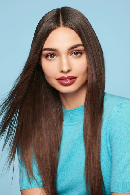 Portrait of a young adult woman with long, straight brown hair, wearing a turquoise sweater, against a light blue backdrop in a studio setting.