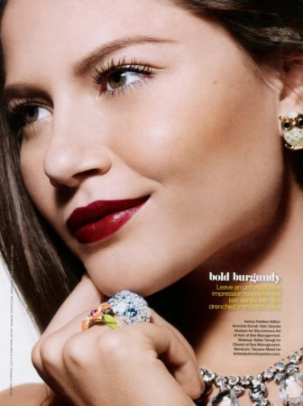Close-up portrait of a woman with dark burgundy lipstick, a floral ring, owl earrings, and a sparkling diamond necklace.