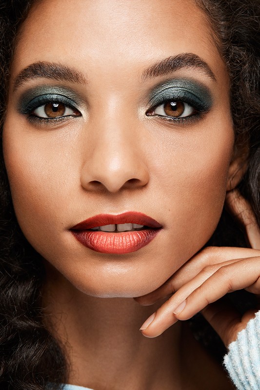 Close-up portrait of a woman with metallic green smokey eye shadow and gradient red lipstick, with her hand near her chin.