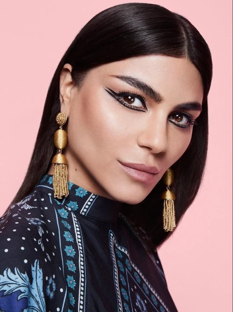 Close-up shot of a woman with dramatic black eyeliner, gold tassel earrings, and a patterned top against a pink background.