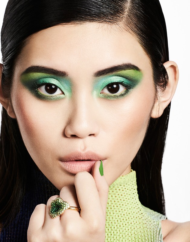 Asian woman close-up with vibrant green makeup, gold ring, and matching green nails posing in a bright photo studio. Beauty shot.