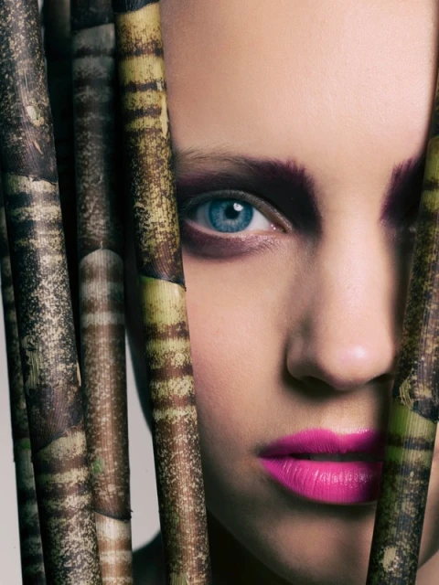 Close-up of a young woman's face with bright blue eyes and vibrant pink lipstick, partially obscured by patterned plant stalks.