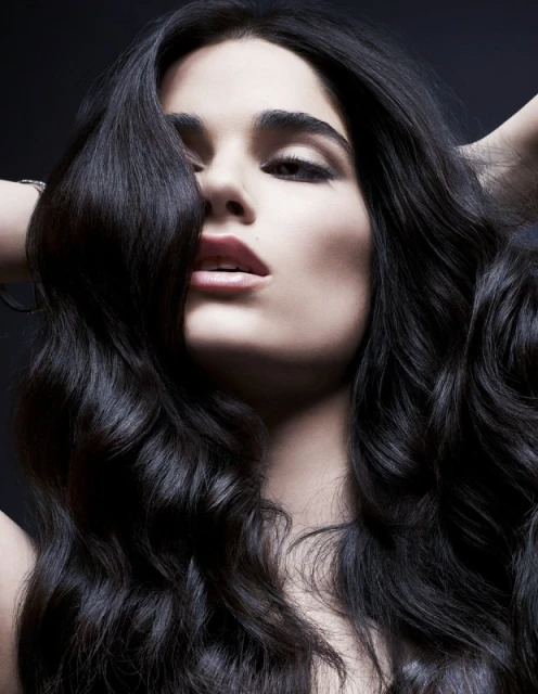 Portrait of a woman with long, dark, wavy hair, hands behind her head, dark eye makeup, and a neutral expression against a dark background.