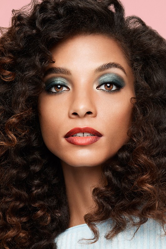 Close-up beauty portrait of a woman with voluminous dark curly hair, shimmering green eyeshadow, and deep red gradient lipstick.