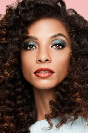 Close-up beauty portrait of a woman with voluminous dark curly hair, shimmering green eyeshadow, and deep red gradient lipstick.
