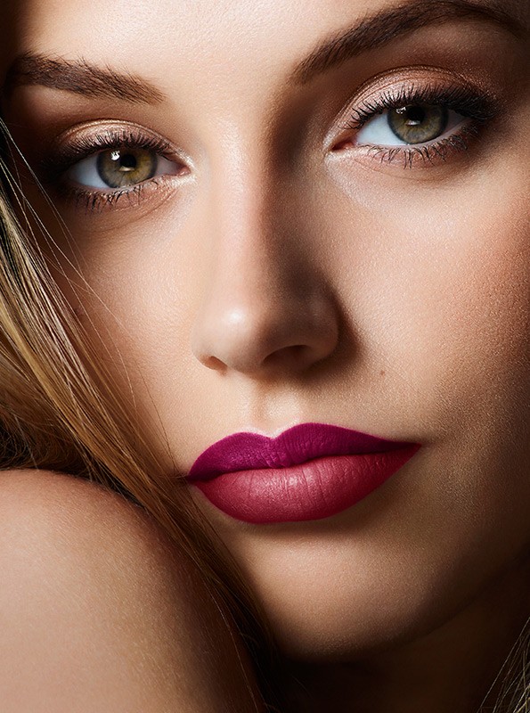 Close-up portrait of a light-skinned young woman with hazel eyes, wearing burgundy lipstick, against a neutral background with soft lighting.