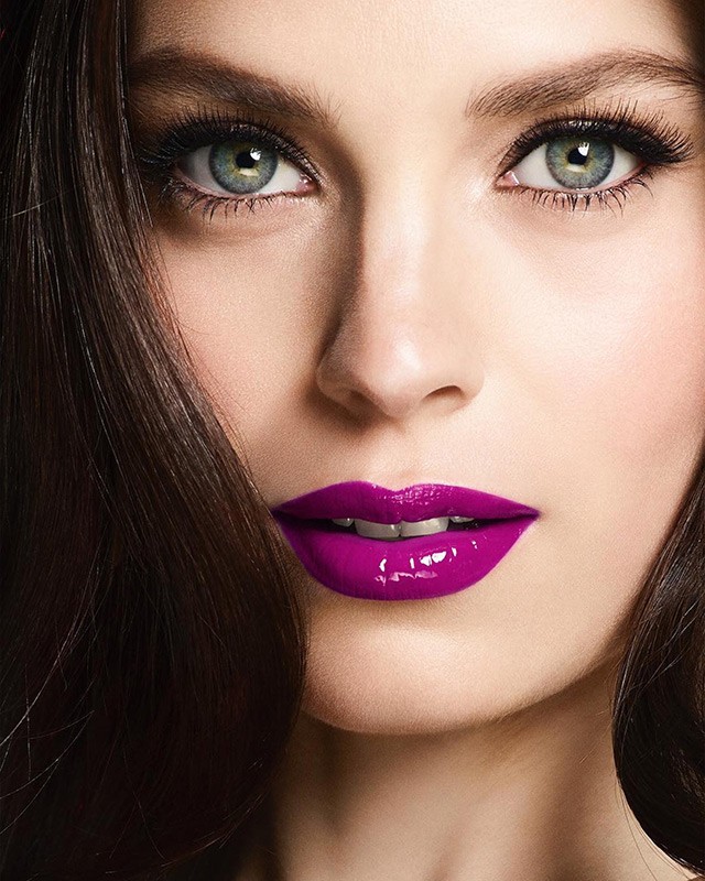 Close-up portrait of an adult woman's face featuring green eyes, long eyelashes, brown hair, and glossy purple lipstick. Beauty and cosmetics.