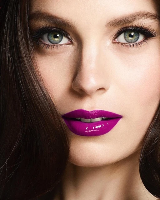 Close-up portrait of an adult woman's face featuring green eyes, long eyelashes, brown hair, and glossy purple lipstick. Beauty and cosmetics.