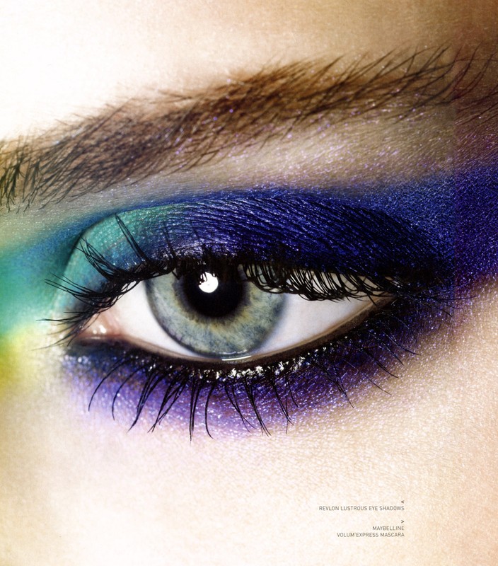 Close-up of a woman's eye with blue and teal eyeshadow and dark mascara, set against a bright, soft background.