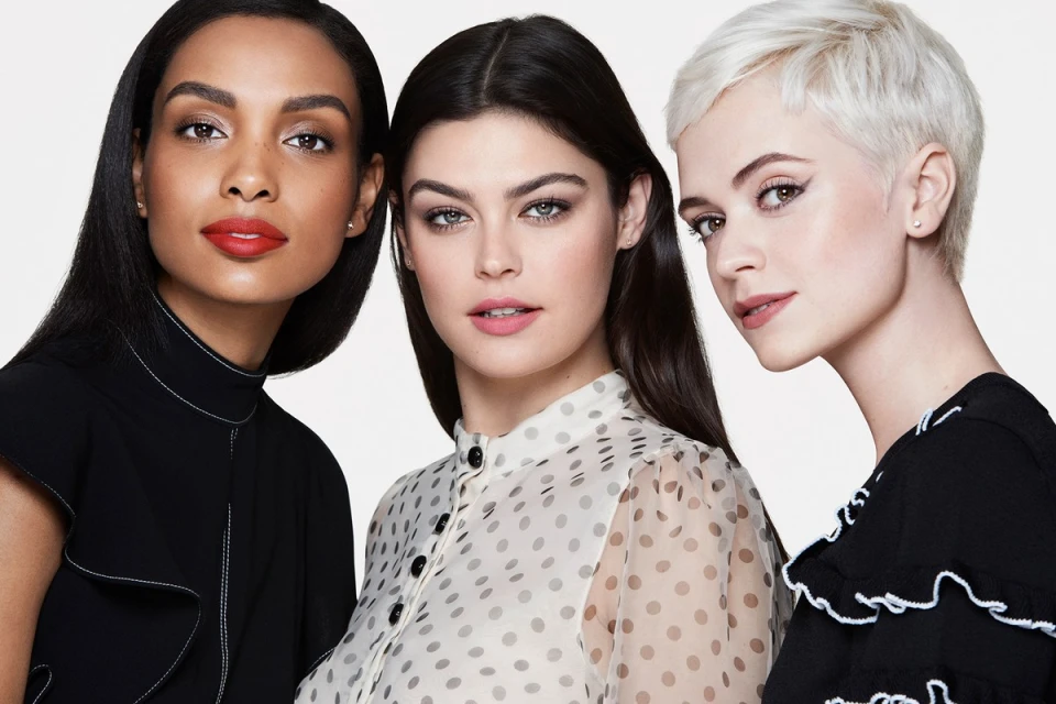 Close-up studio shot of three women with diverse complexions wearing elegant tops and makeup against a clean white backdrop.