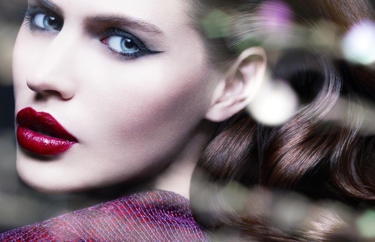 Close-up of a woman with dark hair and bright red lipstick. Her features include a defined eyeliner look and striking blue eyes.