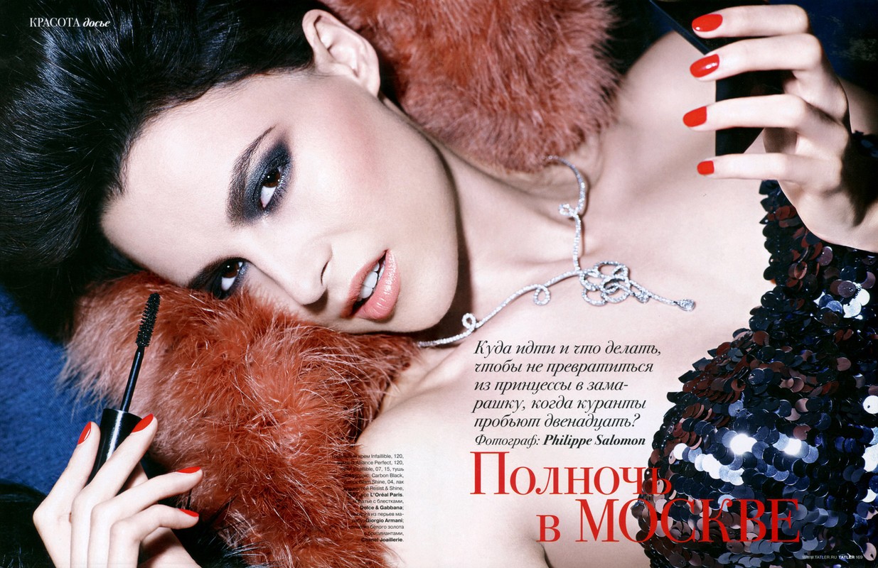 Close-up portrait of a woman wearing dark eye makeup, a sequined dress, and red nail polish while holding a mascara wand.