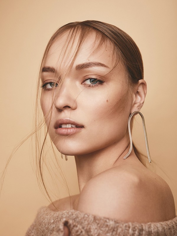 Close-up portrait of a woman with loose strands of hair, wearing statement curved earrings and a textured beige off-the-shoulder top.