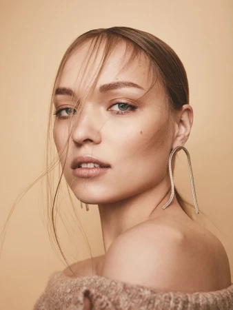 Close-up portrait of a woman with loose strands of hair, wearing statement curved earrings and a textured beige off-the-shoulder top.