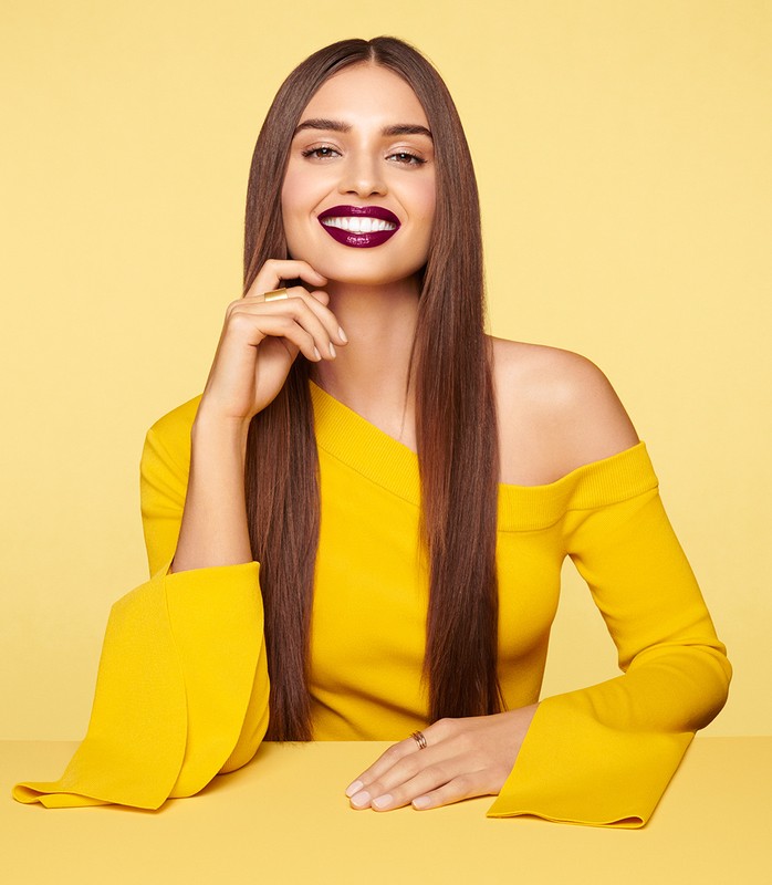 A beautiful woman smiles in a studio shot, wearing a yellow dress, showcasing dark lipstick and hair against a soft yellow background.