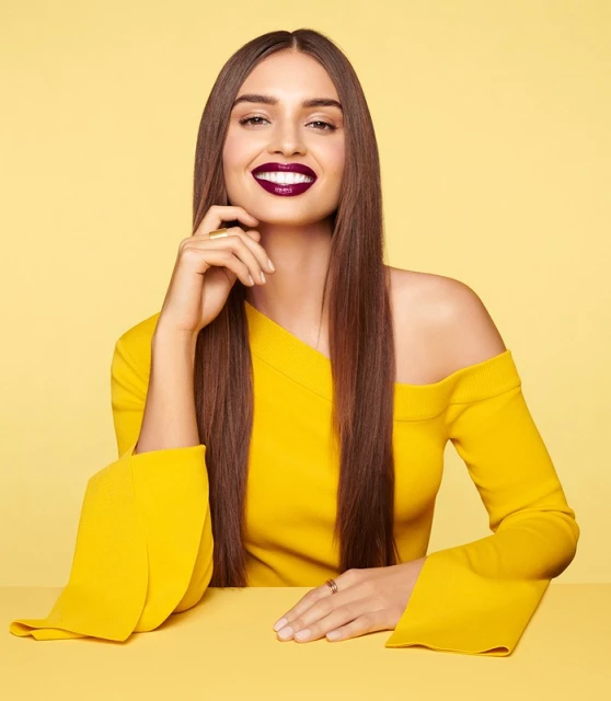 A beautiful woman smiles in a studio shot, wearing a yellow dress, showcasing dark lipstick and hair against a soft yellow background.