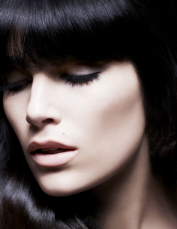 Close-up portrait of a serene adult female with dark wavy hair and closed eyes, emphasizing beauty and dark eye makeup in a studio setting.