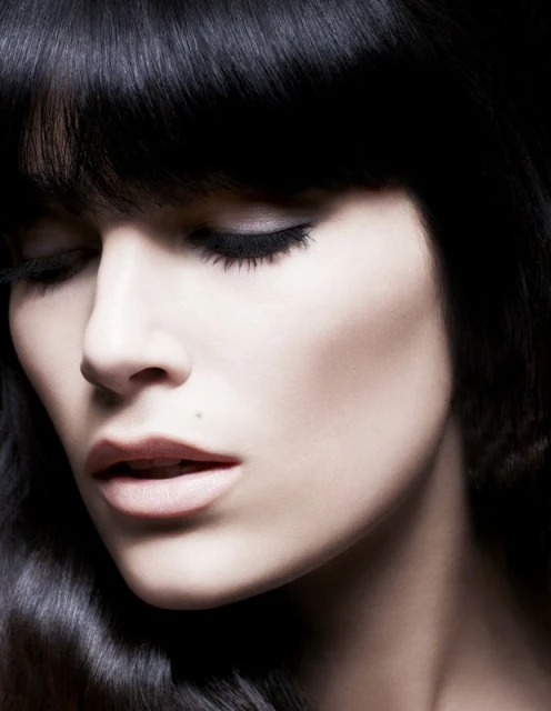Close-up portrait of a serene adult female with dark wavy hair and closed eyes, emphasizing beauty and dark eye makeup in a studio setting.