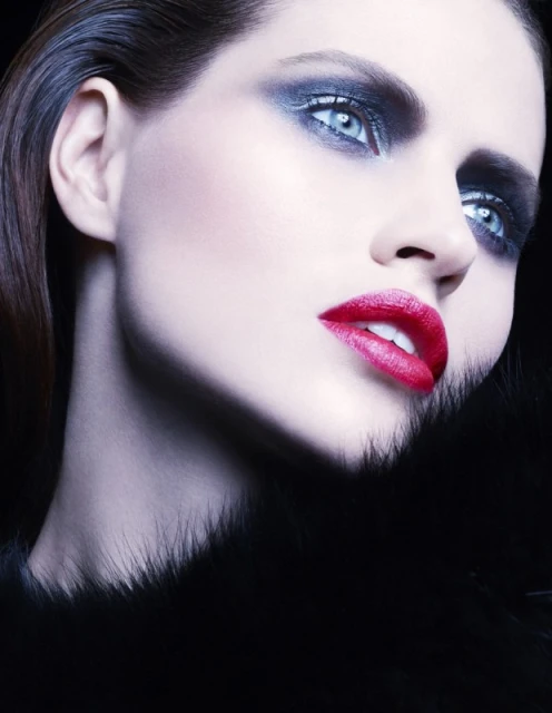 Striking beauty portrait of a woman with bright blue eyes, bold smoky eyeshadow, and red lipstick against a dark background.
