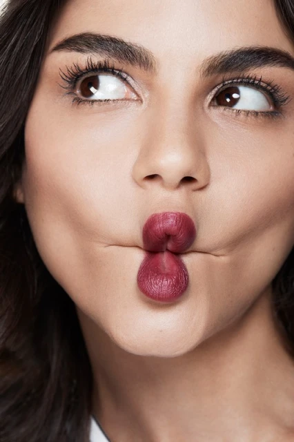 Close-up portrait of a woman with dark hair and brown eyes puckering her lips, set against a plain background in bright, studio lighting.