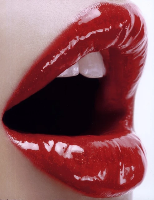 Close-up shot of an adult female's open mouth with glossy red lipstick on the lips and visible white teeth against a white backdrop.