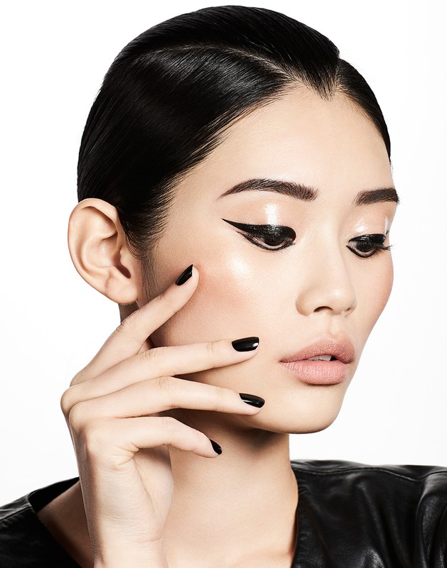 Close-up beauty shot of a young Asian woman with smooth skin, black winged eyeliner, glossy lips, black nails, and black jacket against white.