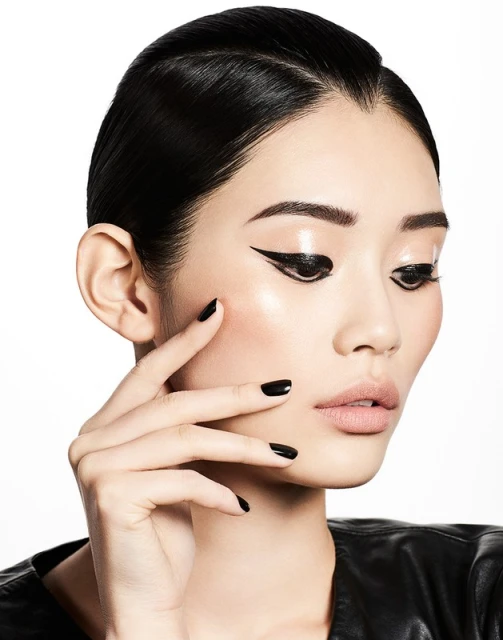 Close-up beauty shot of a young Asian woman with smooth skin, black winged eyeliner, glossy lips, black nails, and black jacket against white.