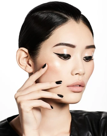 Close-up beauty shot of a young Asian woman with smooth skin, black winged eyeliner, glossy lips, black nails, and black jacket against white.