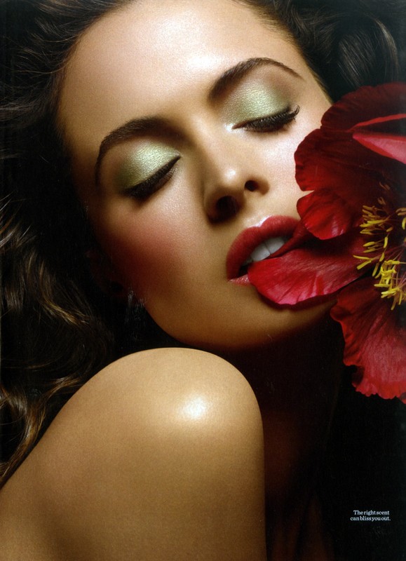 Close-up of a woman with eyes closed, wearing green eyeshadow and red lipstick, with a red flower partially covering her face.