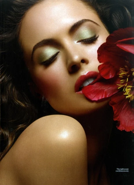Close-up of a woman with eyes closed, wearing green eyeshadow and red lipstick, with a red flower partially covering her face.
