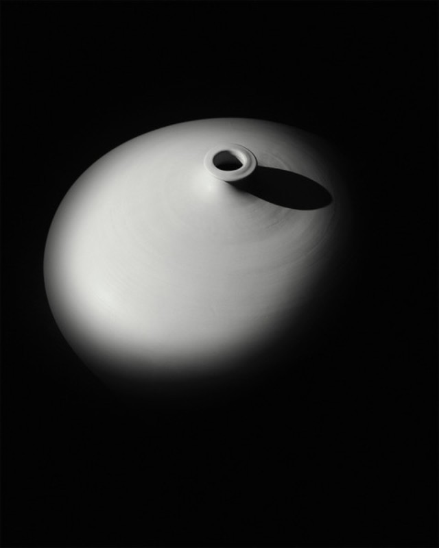 Elevated view of a glossy, cream-colored ceramic vase, illuminated by a single spotlight on a dark background, highlighting its smooth form.