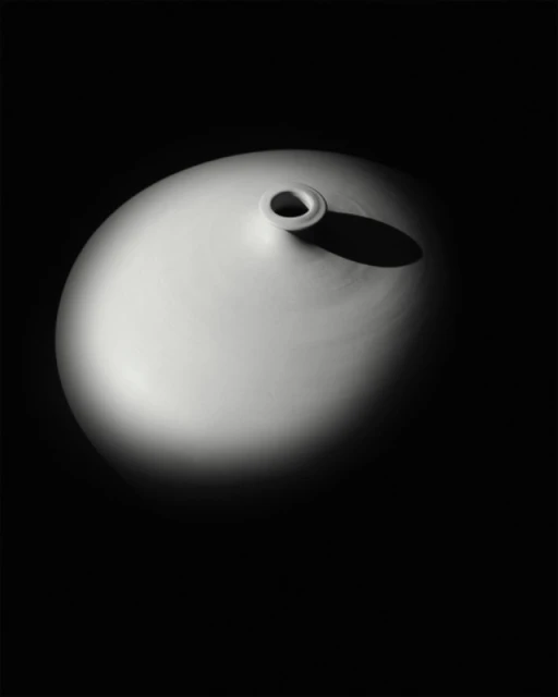 Elevated view of a glossy, cream-colored ceramic vase, illuminated by a single spotlight on a dark background, highlighting its smooth form.