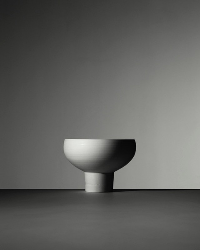 Minimalist still life of a pedestal bowl. The bowl is white ceramic with a matte texture, sitting on a dark surface against a grey backdrop.