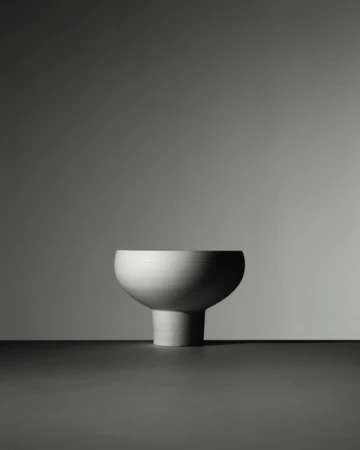 Minimalist still life of a pedestal bowl. The bowl is white ceramic with a matte texture, sitting on a dark surface against a grey backdrop.